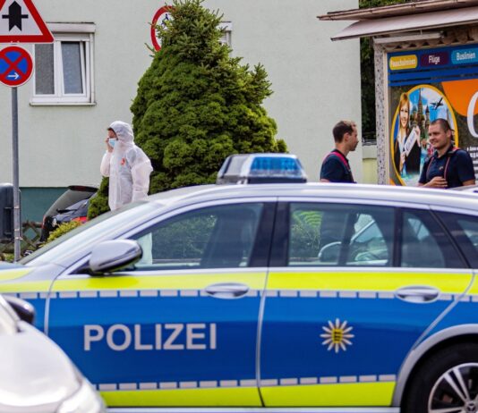 Ein Einsatzfahrzeug der Polizei und im Hintergrund Mitarbeiter der Polizei. Die Spurensicherung ist am Tatort angekommen und sichert die Spuren vor Ort, um den Täter zu finden.