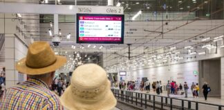 Zwei Fahrgäste blicken voller Erwartung auf die S-Bahn-Linie. Die Station ist unterirdisch und eine Anzeigetafel leuchtet von der Decke. Noch ist keine Bahn zu sehen.