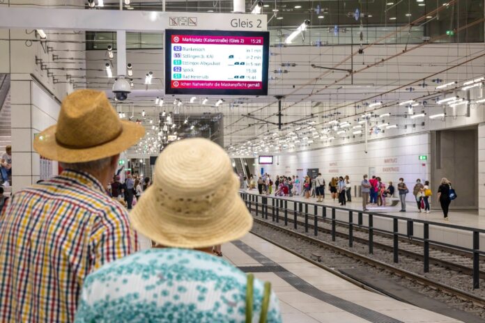 Zwei Fahrgäste blicken voller Erwartung auf die S-Bahn-Linie. Die Station ist unterirdisch und eine Anzeigetafel leuchtet von der Decke. Noch ist keine Bahn zu sehen.