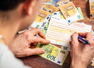 Ein Mann sitzt am Schreibtisch und füllt Felder eines Überweisungsformulars aus. Am Tisch liegen unzählige Euro-Scheine: 50 Euro, 20 Euro und 100 Euro.