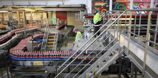 In einer Fabrik werden Coca-Cola-Flaschen auf einem Laufband transportiert. Das Laufband schlängelt sich durch die gesamte Fabrikhalle. Zwei Mitarbeiter prüfen die Abläufe.