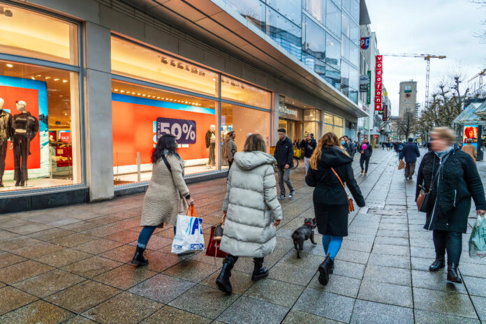 Menschen in einer Einkaufsstraße laufen vor einem Bekleidungsgeschäft vorbei. Sie tragen Tüten und sind gerade auf einer Shopping-Tour in der Innenstadt.