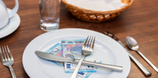 Auf einem Holztisch im Restaurant stehen ein Brotkorb, ein Glas sowie ein Teller. Auf diesem liegen zwei 20-Euro-Geldscheine und überkreuztes Besteck.