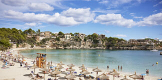 Ein Tag am Strand. Der Sand ist beinahe weiß und gesäumt von Liegen mit Sonnenschirmen und Menschen darunter. Das Meerwasser und der Himmel sind strahlend blau und im Hintergrund erkennt man eine malerische Landschaft. Dies ist eine beliebte Urlaubsinsel der Deutschen.