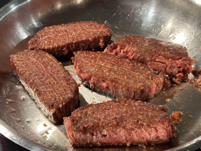 Ein paar vegane Steaks befinden sich in der Pfanne. Einige sind schon gut durchgebraten, andere sind noch in fast rohem Zustand. DIe veganen Steaks sehen fast so aus wie ihre tierischen Originale.