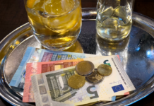 Getränke und Geld auf einem Tablett in einem Restaurant. Ein kleines und ein großes Glas mit Eiswürfeln stehen auf dem silbernen Tablett, auf dem auch ein 20-Euro-Schein, ein 10-Euro-Schein, ein 5-Euro-Schein sowie verschiedene Euro-Münzen liegen.