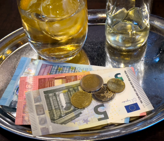 Getränke und Geld auf einem Tablett in einem Restaurant. Ein kleines und ein großes Glas mit Eiswürfeln stehen auf dem silbernen Tablett, auf dem auch ein 20-Euro-Schein, ein 10-Euro-Schein, ein 5-Euro-Schein sowie verschiedene Euro-Münzen liegen.