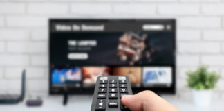 Eine Hand hält eine Fernbedienung und schaltet den Fernseher ein. Es handelt sich um einen Flachbildschirmfernseher. Auf dem Bildschirm kann man verschwommen erkennen, dass offenbar ein Streaming-Dienst durchsucht wird.