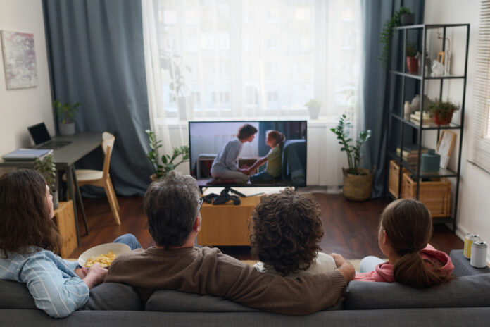 Eine Familie sitzt auf der Couch vor dem Fernseher und schaut fern. Es handelt sich dabei um einen kleinen Flachbildfernseher. Im Hintergrund kann man erkennen, dass es hell ist. Die Gardinen im Wohnzimmer sind zugezogen.