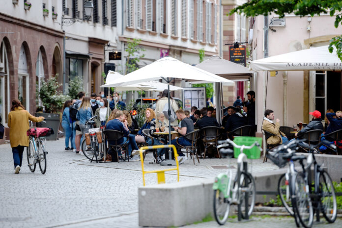 Die Terrasse eines Restaurants im vollen Betrieb mit Gästen. Menschen sitzen an den verschiedenen Tischen unter den weißen Schirmen und essen und trinken etwas.