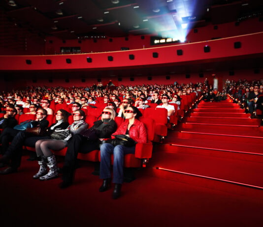 In einem großen Kinosaal sitzen zahlreiche Besucher auf roten Kinosesseln und haben 3D-Brillen auf. Sie blicken gespannt auf die Leinwand vor ihnen, während der Filmprojektor im Hintergrund läuft.