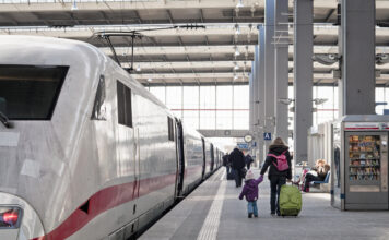 An einem Bahnhof, wo zurzeit wenig los ist, steht ein weißer ICE hält an einer Station. Eine Mutter mit Koffer und ihrem Kind läuft das Gleis entlang, um am richtigen Abschnitt des Gleises in den Wagon des Zuges einzusteigen.