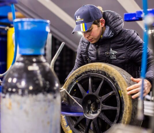 Ein Mitarbeiter in einer Kfz-Werkstatt hält einen Reifen in der Hand um ihn zu wechseln. Er arbeitet in einem Betrieb mit Fahrzeugen.