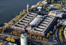 Luftaufnahme einer großen Industriehalle umgeben von mehreren Grünflächen. Links von der Fabrik liegt der Rhein. An das Industriegelände grenzen andere Gebäude.