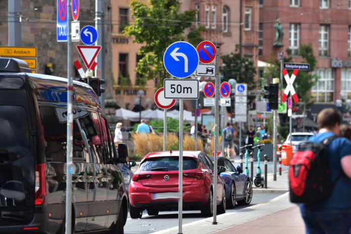 In einer belebten, viel befahrenen Straße in der Innenstadt, von Menschen und Autos durchströmt, sind aber rechten Straßenrand unzählige verschiedene Verkehrsschilder und Straßenschilder angebracht.