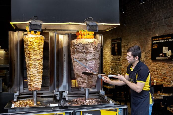 Ein Mann schneidet Döner-Fleisch. Der Mitarbeiter steht in einem Döner-Imbiss und bereitet die beliebte türkische Mahlzeit für die Kunden zu. Den Döner gibt es im Fladenbrot mit Kraut und Fleisch.