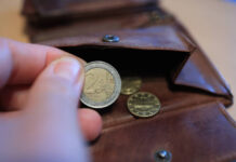 Eine Hand hält eine 2-Euro-Münze zwischen Zeigefinger und Daumen über einem Geldbeutel. In dem leicht geöffnetem Portemonnaie aus braunem Leder liegt noch eine 10-Cent- und eine 20-Cent-Münze.
