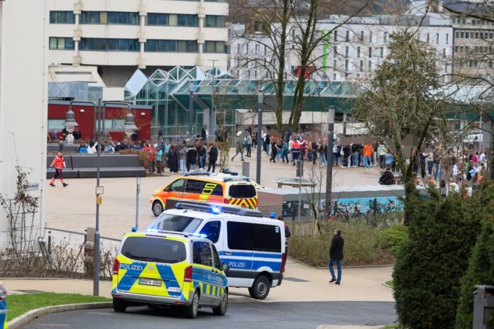 Zwei Polizeiwagen mit Blaulicht und ein Feuerwehr-Auto kommen zu einem Einsatz an einem großen öffentlichen Platz, umringt von mehreren großen Gebäuden. Rechts bildet sich eine größere Menschenmasse.