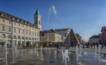Der Karlsruher Marktplatz und die gesamte Innenstadt an einem heißen Tag mit dem Brunnen. Die Menschen flanieren über den Platz und versammeln sich am Wasser.