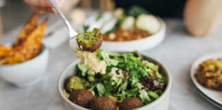 Eine Person isst eine Falafel-Bowl mit Gemüse und Salat. Im Hintergrund stehen gesunde vegane und vegetarische Snacks auf dem Tisch.