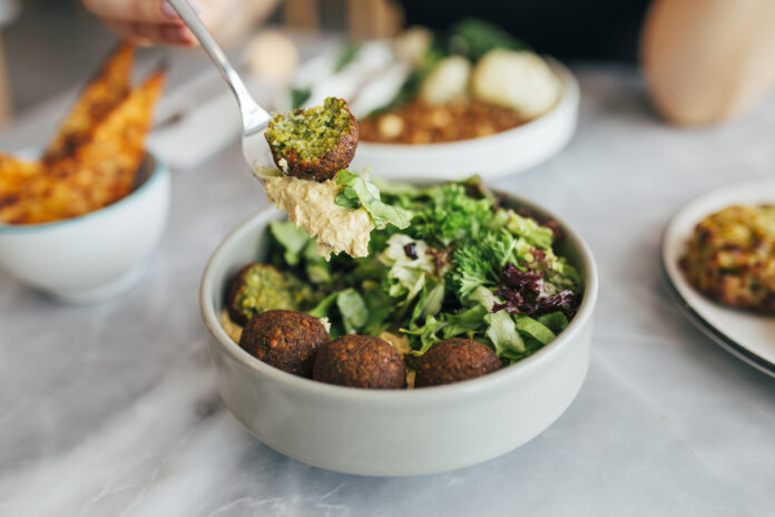 Eine Person isst eine Falafel-Bowl mit Gemüse und Salat. Im Hintergrund stehen gesunde vegane und vegetarische Snacks auf dem Tisch.