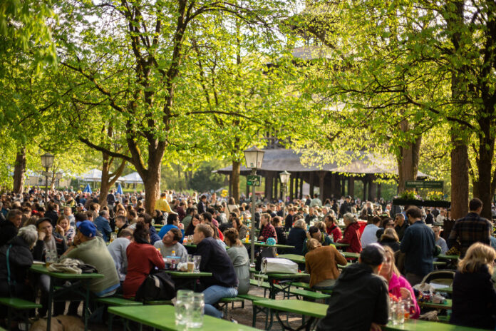 Viele Menschen sitzen auf grünen Bänken in einem Biergarten mit großen grünen Bäumen.