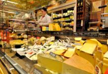 Eine Verkäuferin befindet sich hinter der Käsetheke und verkauft Käse. In der Auslage gibt es viele verschiedene Sorten, unter denen sich die Kunden ihre Wunschprodukte aussuchen können.