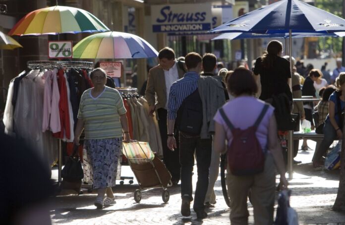 Viele Passanten schlendern durch die Sonne, einige halten einen bunten Sonnenschirm in der Hand. Sie kaufen in einigen Geschäften der Innenstadt ein und betrachten die Auslagen der Händler. Ab dem 1. Juli erwarten die Bürgerinnen und Bürger neue Pflichten.