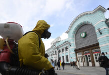 Ein Mann in einem kompletten Schutzanzug mit einer Gasmaske desinfiziert mit einem Gerät eine Bahnhofsstation. Im Hintergrund laufen einige Leute in und aus dem Bahnhof. In einer deutschen Großstadt breitet sich ein Virus aus.