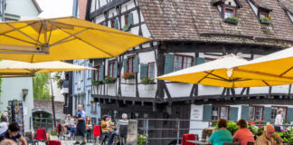 Ein Blick auf das schiefste Hotel der Welt. Es befindet sich in Ulm und ist ein altes Fachwerkhaus. Vor dem Hotel ist ein Café und mehrere Gäste sitzen unter großen Sonnenschirmen und trinken Kaffee.