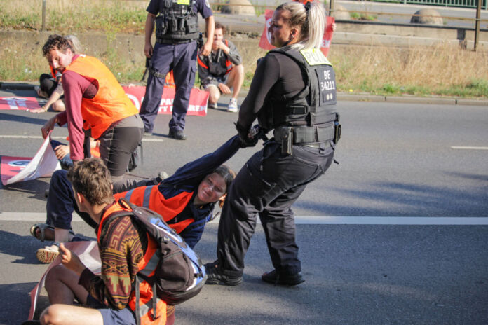 Mehrere in Warnwesten gekleidete, an die Straßen geklebte Klimaaktivisten der Gruppierung "Letzte Generation" werden von der Polizei unter Anwendung eines Griffes von der Fahrbahn gelöst und weggebracht, um die Straße wieder frei für Autos zu machen.