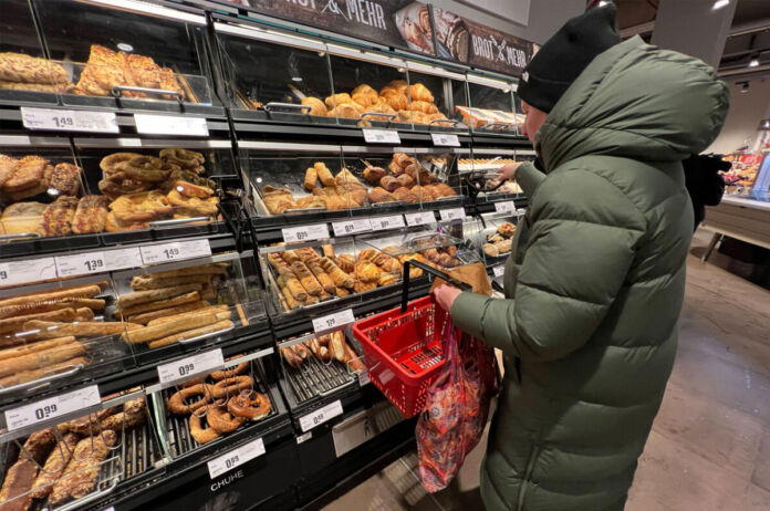 Ein Kunde steht vor einem Backregal in einem Supermarkt oder Discounter. Er schaut sich die Auslagen an, die vollen Brötchen, Brote und Baguette sind. Er trägt einen Einkaufskorb bei sich und überlegt offenbar, was er einkaufen will.