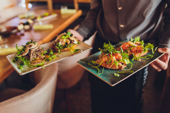 Ein Kellner trägt zwei köstlich aussehende Fleischplatten, ohne dass man dabei seinen Kopf erkennt. Fokus liegt auf den Tellern mit dem Essen. Salate schmücken das Ganze als Beilage. Für diese Gerichte verlangt das Restaurant wahrscheinlich überdurchschnittliche Preise.