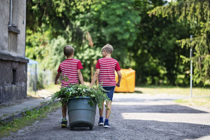 Zwei Jungs ziehen einen großen Eimer mit Kompost hinter sich her. Grüne Pflanzen ragen aus dem Behälter. Die Kinder sind blond.