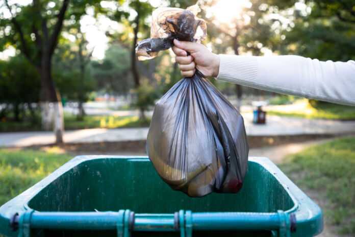 Eine Person wirft einen Müllsack in eine grüne Mülltonne. Es handelt sich vermutlich um Hausmüll oder gemischten Müll. Der Müllsack ist schwarz und teilweise durchscheinend.