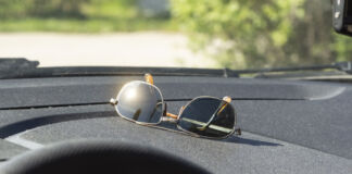 Eine Sonnenbrille liegt unter der Windschutzscheibe im Auto im Cockpit. Die Sonne scheint darauf und ein Glas blendet die Sonne drastisch zurück.