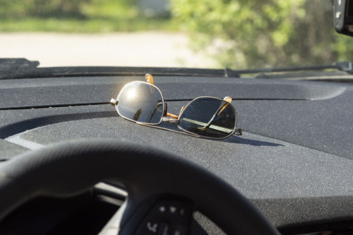 Eine Sonnenbrille liegt unter der Windschutzscheibe im Auto im Cockpit. Die Sonne scheint darauf und ein Glas blendet die Sonne drastisch zurück.