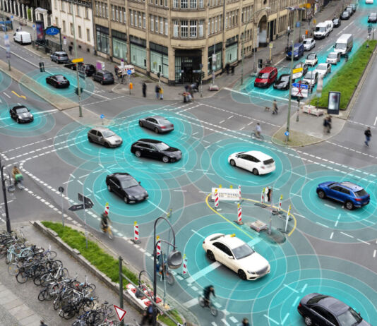 Eine belebte Straßenkreuzung in der Stadt mit regem Verkehr. Verschiedene Autos, Fahrradfahrer und Fußgänger bewegen sich. Um die Autos sind blaue Kreise dargestellt, um zu veranschaulichen, dass sie moderne Sensoren für räumliches Bewusstsein eingebaut haben. Es könnten neue E-Autos oder selbstfahrende Fahrzeuge sein.