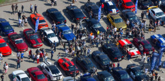 Auf einem riesig großen Parkplatz stehen viele Ford Mustangs unterschiedlicher Jahre. Eine Menschenmasse läuft zwischen den einzelnen Modellen entlang und schaut sich jedes an.