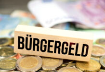 Bargeld, vor allem zwei Euro Münzen und verschiedene Geldscheine, liegen auf einem Tisch. Darauf liegt ein Schild aus Holz mit der Aufschrift Bürgergeld