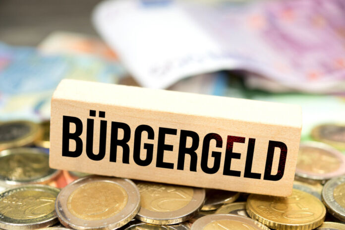 Bargeld, vor allem zwei Euro Münzen und verschiedene Geldscheine, liegen auf einem Tisch. Darauf steht ein Schild aus Holz mit der Aufschrift Bürgergeld. Nun kommt eine neue Kostenpflicht für Bürgergeld-Empfänger.