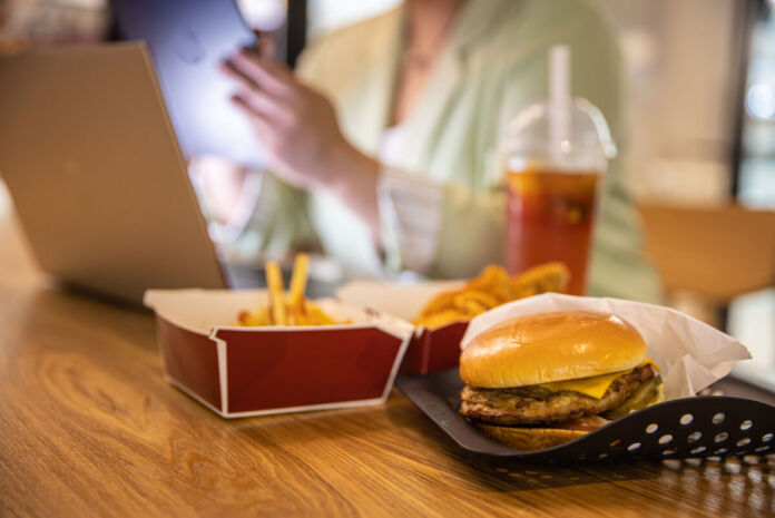 Eine Frau sitzt an einem Tisch, auf dem ein Laptop steht. Im Vordergrund sieht man eine Schachtel mit Pommes und ein Tablett, auf dem ein Burger liegt.