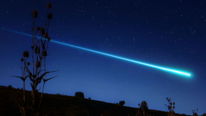 Ein Meteor fliegt hoch am Himmel. Er hinterlässt einen langen und leuchtenden Schweif. Vorne im Bild sind einige Bäume zu sehen.