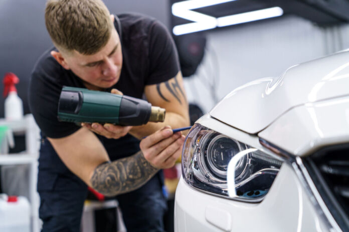 Ein Mitarbeiter einer Werkstatt klebt etwas mit einem Heißkleber und Werkzeug an einem Scheinwerfer eines Autos fest. Das Fahrzeug ist weiß und der Mitarbeiter konzentriert sich auf die Arbeit.