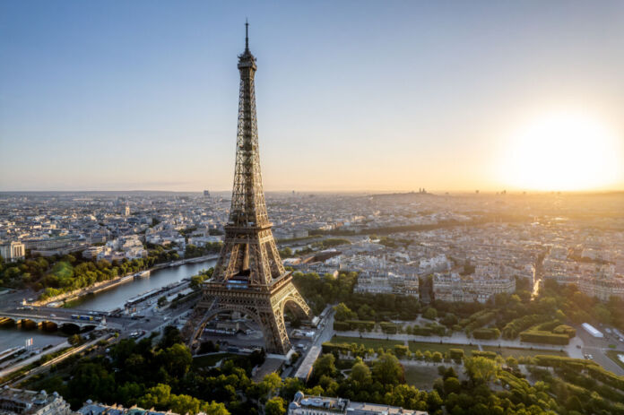 Die Stadt Paris mit präsentem Eiffelturm im Morgengrauen. Am wolkenlosen Himmel geht die Sonne auf. Der Horizont reicht weit über die Skyline der Hauptstadt von Frankreich.