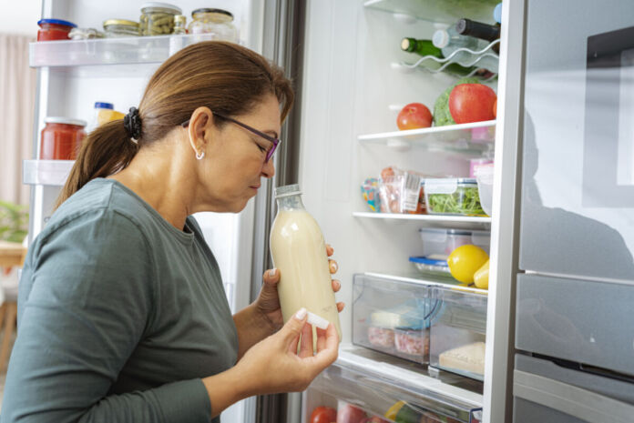 Eine Frau steht vor dem geöffneten Kühlschrank und riecht an einer Flasche Milch. Ihr Blick zeigt, dass sie sich ekelt, denn es handelt sich um abgelaufene Milch.