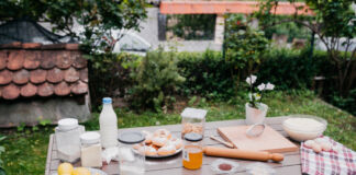 Ein Tisch in einem grünen Garten. Auf dem Tisch befinden sich verschiedene Lebensmittel wie Zitronen und Orangen. Aber auch ein Glas Milch und eine Schüssel mit Mehl sind dabei. Zusätzlich stehen Dosen mit Zucker auf dem Tisch.