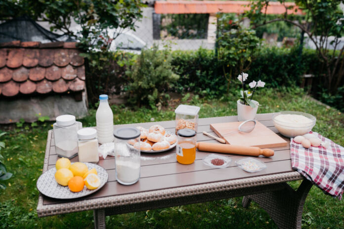 Ein Tisch in einem grünen Garten. Auf dem Tisch befinden sich verschiedene Lebensmittel wie Zitronen und Orangen. Aber auch ein Glas Milch und eine Schüssel mit Mehl sind dabei. Zusätzlich stehen Dosen mit Zucker auf dem Tisch.