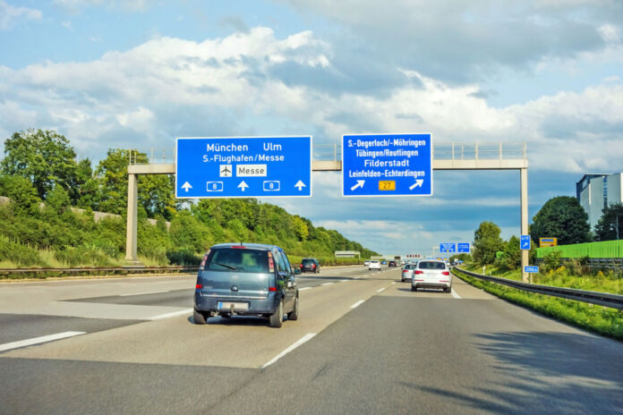 Eine sonnige Autobahn in Deutschland im Sommer, auf der zahlreiche Autos und Kleinwagen fahren. Über ihnen hängen einige verkehrsweisende Streckenschilder.