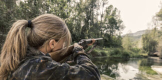 Eine Frau mit einer braun grünen Jacke und einem Zopf ist von hinten zu sehen, während sie mit einem Gewehr im Wald steht. Die Waffe ist auf ein Tier gerichtet, welches sie jagen möchte.